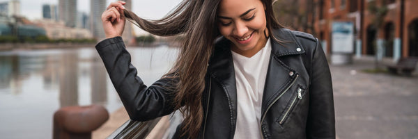 Smiling woman in a black leather jacket standing by a riverfront with city buildings in the background, featured as the cover photo for a Leather Jacket Buying Guide.