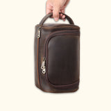 A person holding the High Plains Leather Toiletry Bag by its sturdy leather handle, displaying its durable full-grain leather construction and practical front zipper compartment.